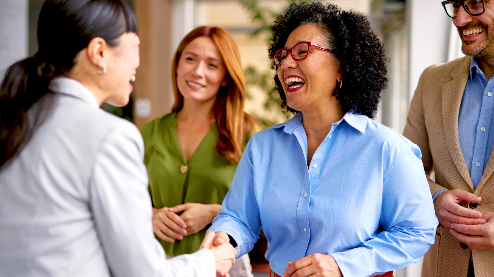 A Black woman and Asian woman shaking hands during a job interview with a white man and redheaded woman happily looking on