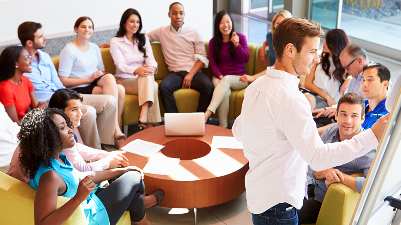 A business meeting with a white man writing on a whiteboard while surrounded by a diverse team paying attention